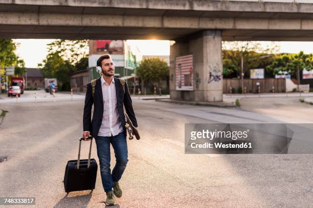 young man on the move with skateboard, rolling suitcase and headphones - mala de rodinhas - fotografias e filmes do acervo