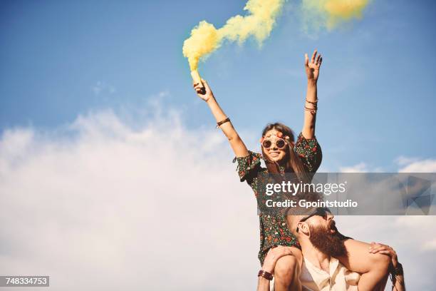 young boho woman holding yellow smoke flare on boyfriends shoulders at festival - festival a stock-fotos und bilder