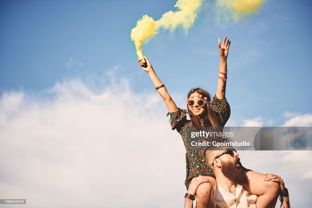 Young boho woman holding yellow smoke flare on boyfriends shoulders at festival
