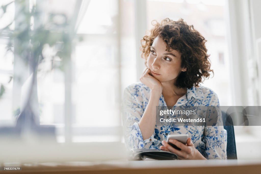 Businesswoman in office with smartphone and diary, looking worried