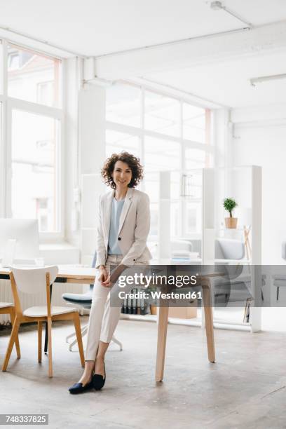 businesswoman in office sitting on desk, looking confident - female suit stockfoto's en -beelden