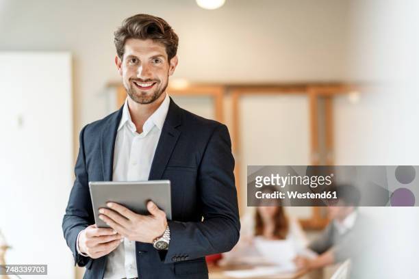 portrait of confident businessman holding tablet with a meeting in background - businesswear stock pictures, royalty-free photos & images