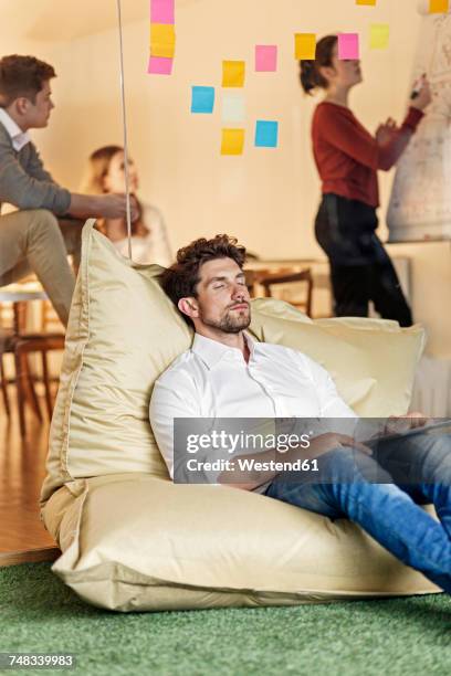 relaxed man in office sitting in bean bag with meeting in background - bean bag stock-fotos und bilder