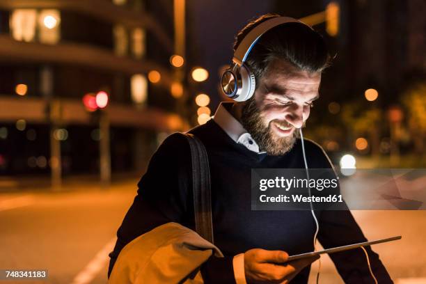 smiling young man with tablet and headphones on urban street at night - ipad kopfhörer stock-fotos und bilder