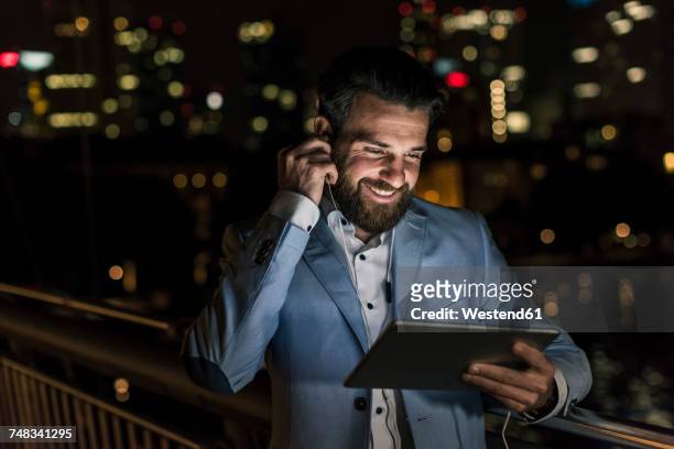 smiling young man with tablet and earphone on urban bridge at night - man business hipster dark smile stock pictures, royalty-free photos & images