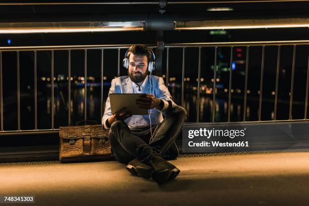 smiling young man with tablet and headphones sitting on bridge at night - man business hipster dark smile stock pictures, royalty-free photos & images