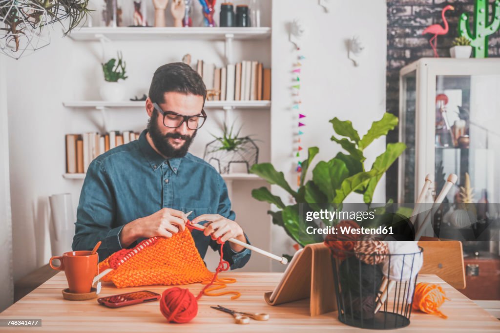 Bearded man knitting at home using tablet for watching online tutorial