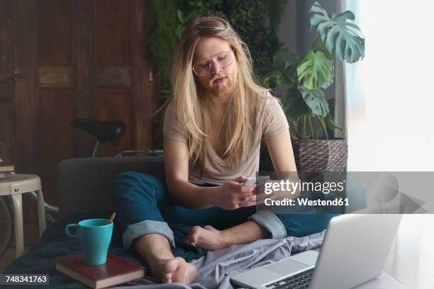 man with long hair and beard sitting on sofa bed looking at smartphone - casa real española fotografías e imágenes de stock