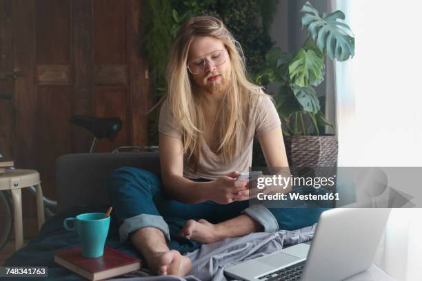 man with long hair and beard sitting on sofa bed looking at smartphone - long blonde hair stock pictures, royalty-free photos & images