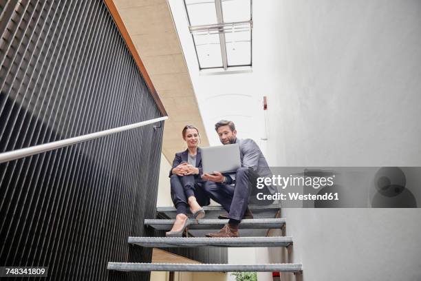 businessman and businesswoman sitting on stairs in office sharing laptop - man stairs sitting stock-fotos und bilder