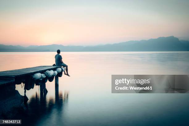 italy, lazise, man sitting on jetty looking at lake garda - loneliness concept stockfoto's en -beelden