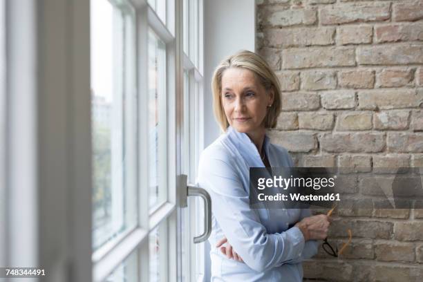 mature businesswoman standing at window, looking worried - teleurstelling stockfoto's en -beelden
