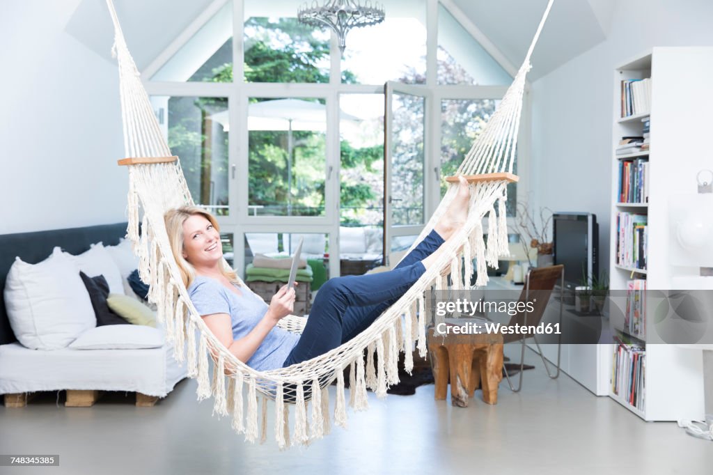 Smiling blond woman with tablet relaxing in hammock in the living room