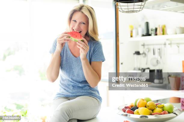 mature woman sitting in kitchen, eating water melon - mature woman in water stock pictures, royalty-free photos & images