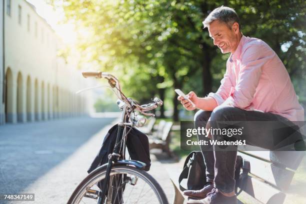 smiling man with bicycle checking the phone on a park bench - commuter man europe bike stock-fotos und bilder