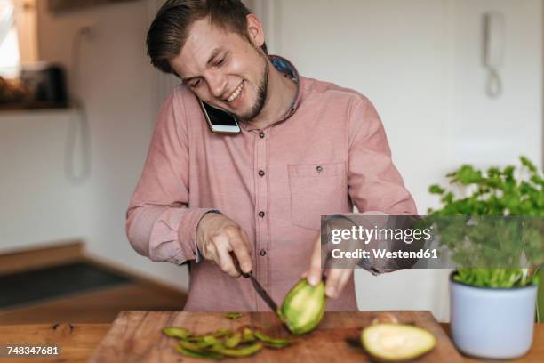 smiling man on the phone chopping acocado in the kitchen - tête penchée photos et images de collection