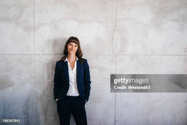 businesswoman leaning against office wall with hands in pockets - three quarter length stock pictures, royalty-free photos & images