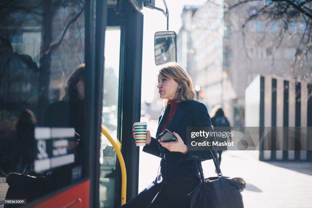 Woman with disposable cup and smart phone standing by bus in city on sunny day