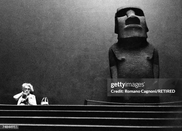 An elderly woman sits reading by a large stone statue, or moai from Easter Island on display at the British Museum, London, 1967.