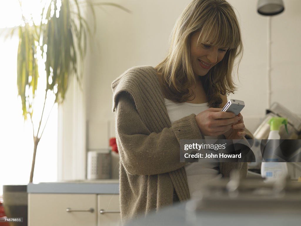Young woman standing at kitchen sink, using cell phone, smiling