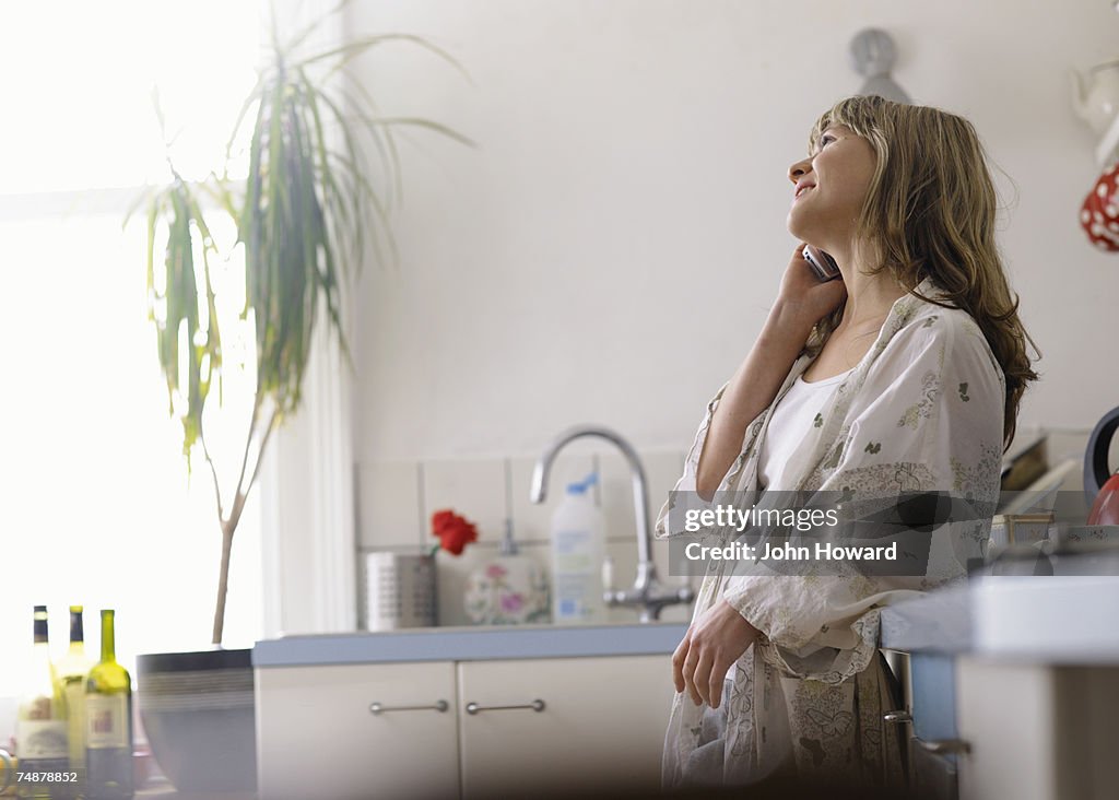 Young woman leaning on kitchen sink, using cell phone, smiling