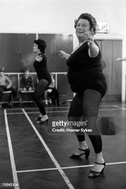 An overweight grandmother wearing high heel shoes in a keep fit dance class while exercising to pop music, Essex, circa 1980. Her daughter and...