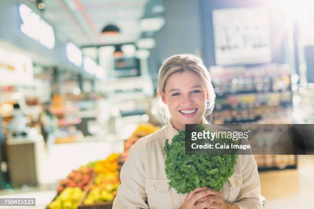 portrait smiling young woman holding bunch of kale in grocery store market - kale bunch stock pictures, royalty-free photos & images