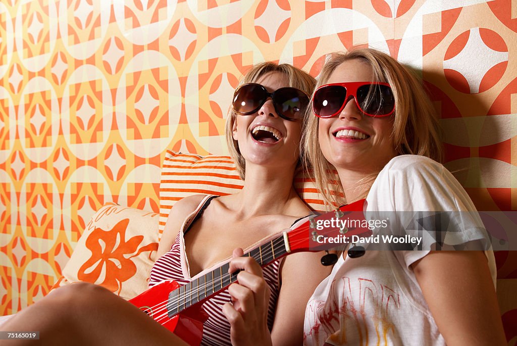 Two young women wearing sunglasses, sitting on bed, one playing guitar, laughing