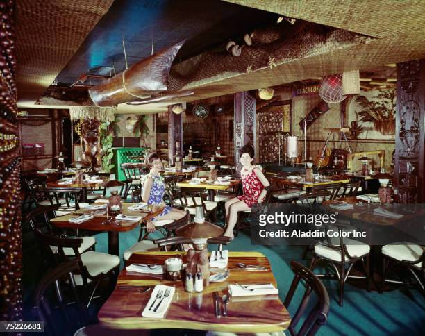 Two Beachcomber Hotel staff members, both dressed in Hawaiian-style, flower-print dresses, sit in the hotel's Polynesian themed restaurant,...