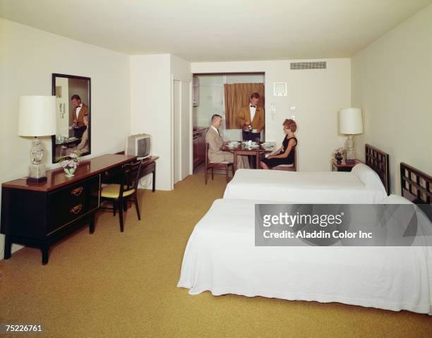 Couple, the man in a suit and the woman in a dress, sit at a table in their room at the Carousel hotel as a uniformed room service attendant serves...