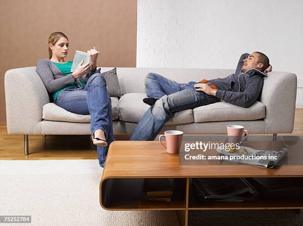 young woman reading book and young man listening to mp3 player on sofa - coffee table reading mug stock pictures, royalty-free photos & images