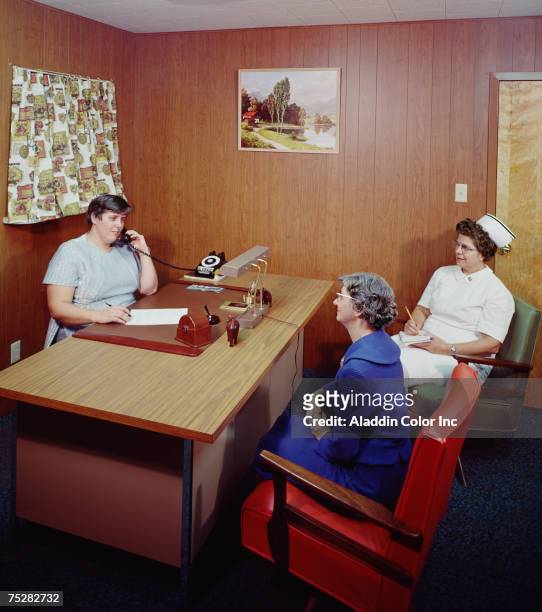 An elderly woman sits with a nurse in a wood-panelled office at the Van Rensselaer Manor Nursing Home, while another staff member makes a telephone...