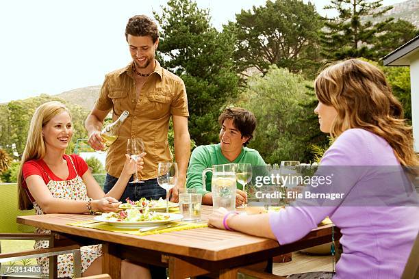 2 young smiling couples sitting at garden table, man pouring wine - four people foto e immagini stock