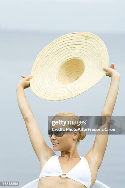 young woman wearing sunglasses, holding up sunhat, head and shoulders - strohhut stock-fotos und bilder