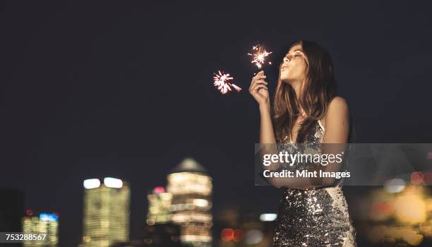 a young woman in a sequined dress dancing on a rooftop at night holding a party sparkler. - avondkledij stockfoto's en -beelden