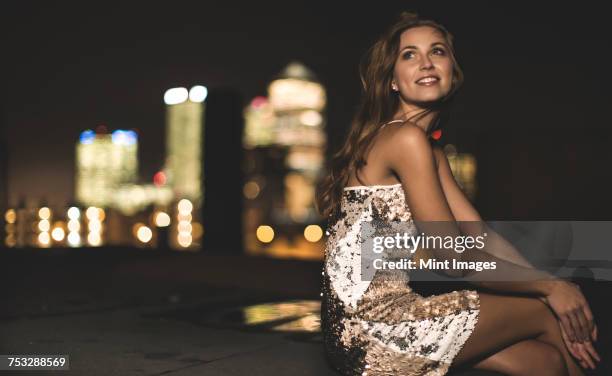 a young woman in a sequined party dress sitting on a rooftop at night. - vestido de fiesta fotografías e imágenes de stock