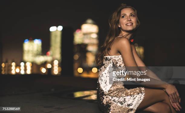 a young woman in a sequined party dress sitting on a rooftop at night. - vestito da sera femminile foto e immagini stock