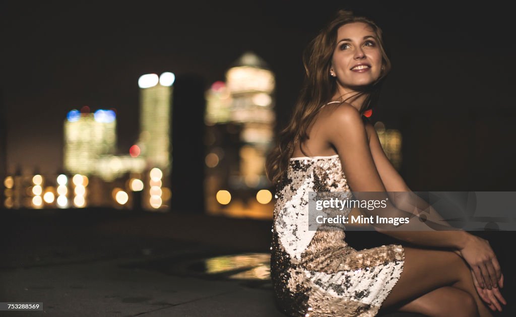 A young woman in a sequined party dress sitting on a rooftop at night.