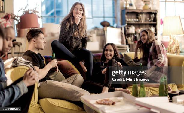 four young women and young man sitting on a sofa, smiling, pizza and beer bottles on coffee table. - beer friends stockfoto's en -beelden