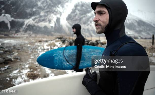 two surfers wearing wetsuits and carrying surfboards standing on a beach with mountains behind. - surfer wetsuit stockfoto's en -beelden