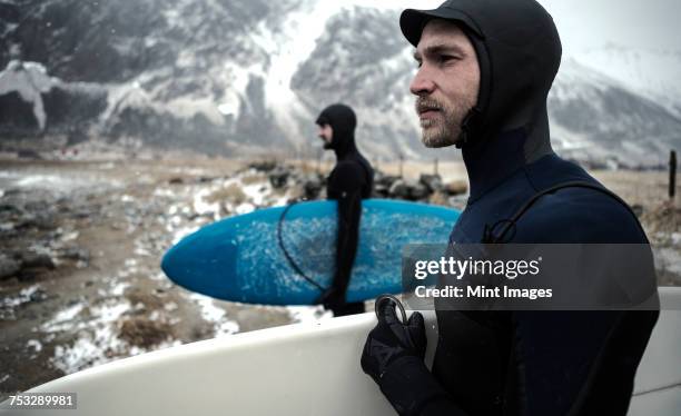 two surfers wearing wetsuits and carrying surfboards standing on a beach with mountains behind. - surfer wetsuit stock pictures, royalty-free photos & images