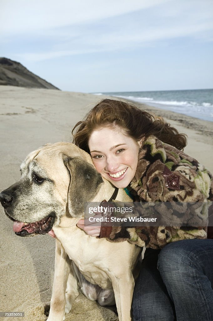 Teenage girl (16-18) crouching by dog on beach, smiling, portrait