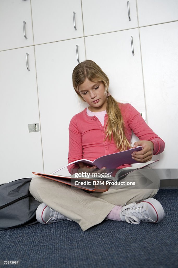 Girl (10-11) sitting in front of lockers, reading book