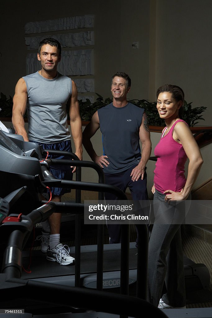 Three people standing on treadmill in gym, smiling, portrait