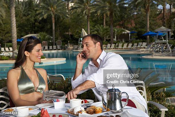 couple sitting by table at poolside, man using mobile phone - las vegas pool stock pictures, royalty-free photos & images