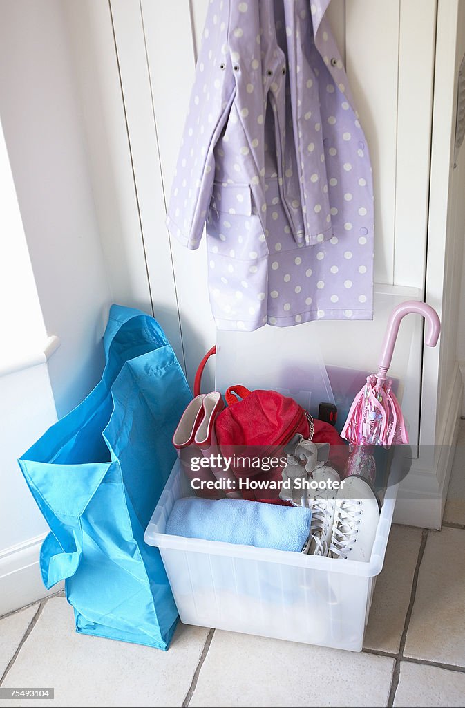 White plastic box containing shoes, boots, umbrella, towel, with blue bag next to it, and mauve raincoat with hanging on back of door