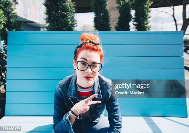 portrait of redhead young woman gesturing peace sign while sitting on bench - gestikulieren stock-fotos und bilder