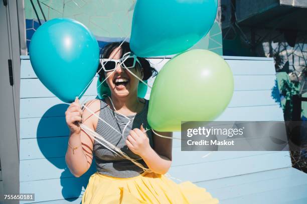 happy young woman holding balloons while sitting on bench - blue sunglasses stock pictures, royalty-free photos & images