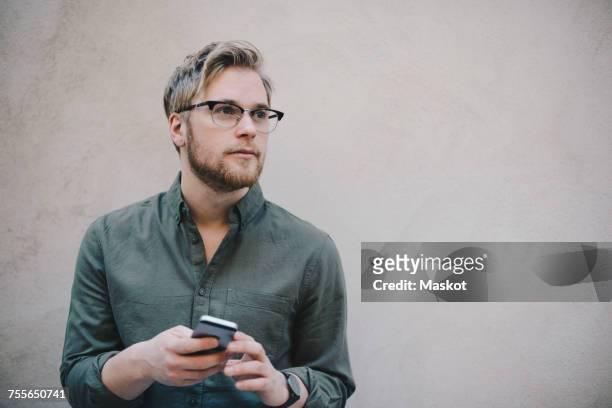 thoughtful male computer programmer holding smart phone against beige wall in office - beige shirt stock pictures, royalty-free photos & images