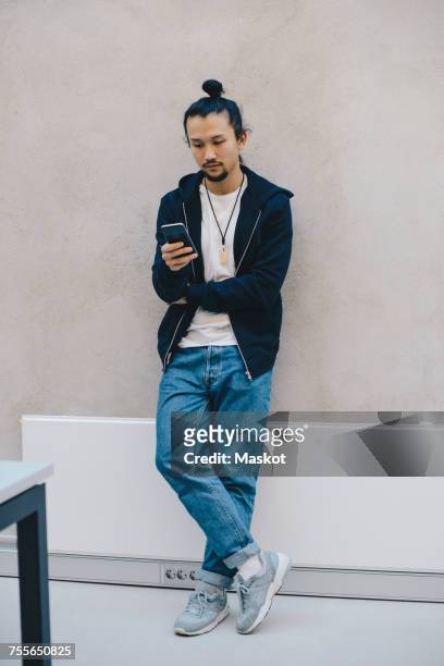 full length of male computer programmer using smart phone while leaning on beige wall in office - casual male standing stock-fotos und bilder